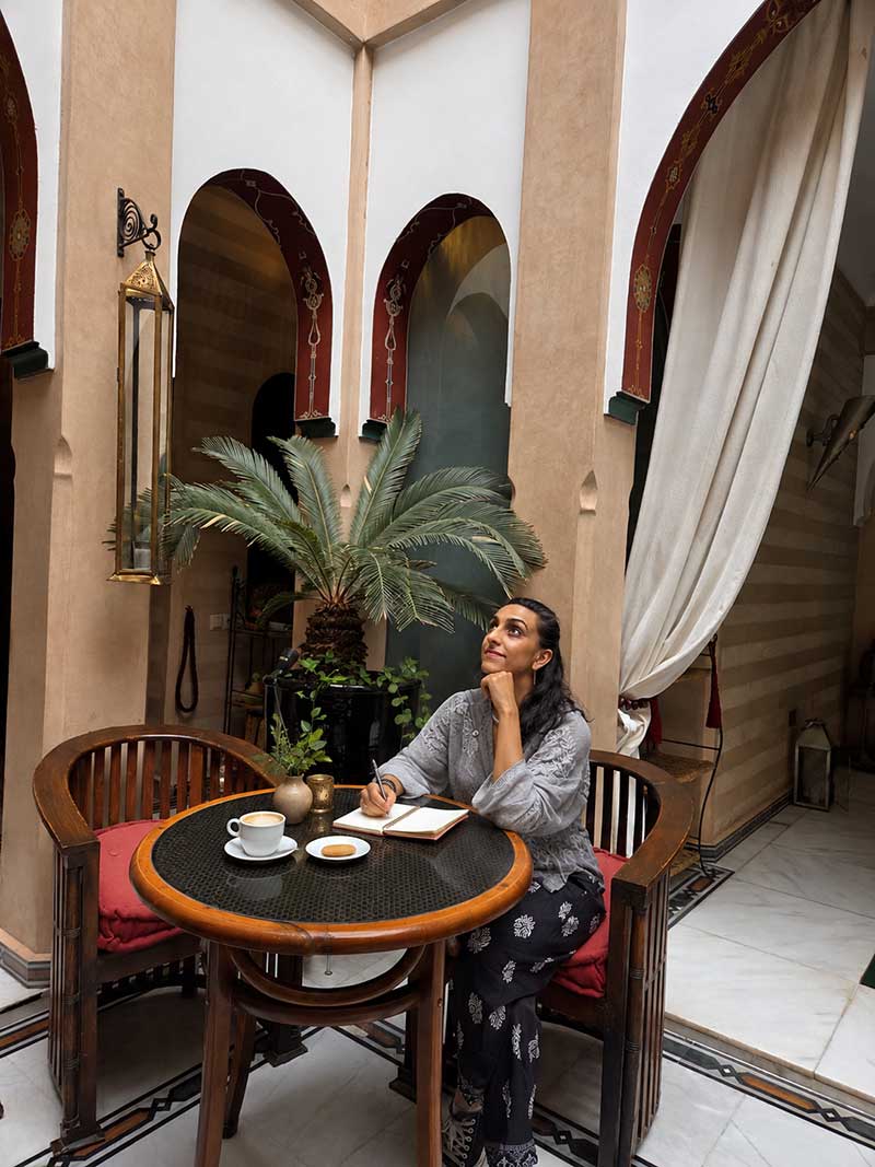 Dr Gauri Chauhan at a café table, pen in hand, looking up from an open notebook.
